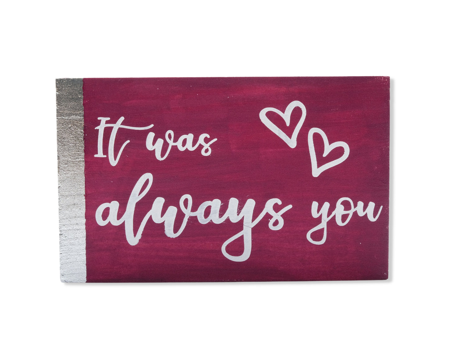Small freestanding rectangular wooden sign facing straight on. Magenta color, silver vertical border on left side only. White painted message, It was always you, with two heart outlines in top right corner. Studio style photo on white background.