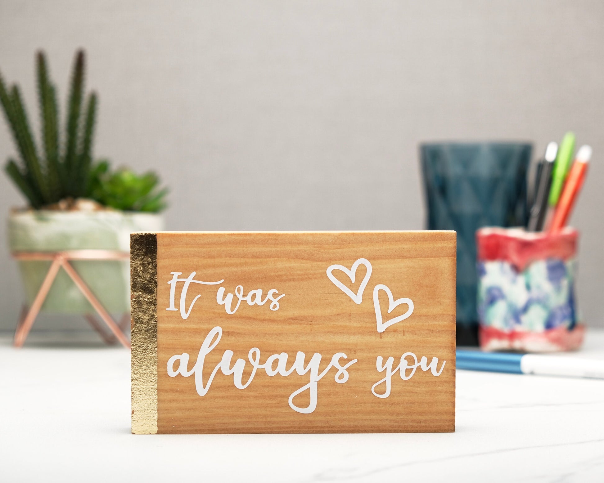Small freestanding rectangular wooden sign facing straight on. Natural wood with gold vertical border on left side. White painted message, It was always you, with two heart outlines top right corner. Plant and pen pot out of focus in background.