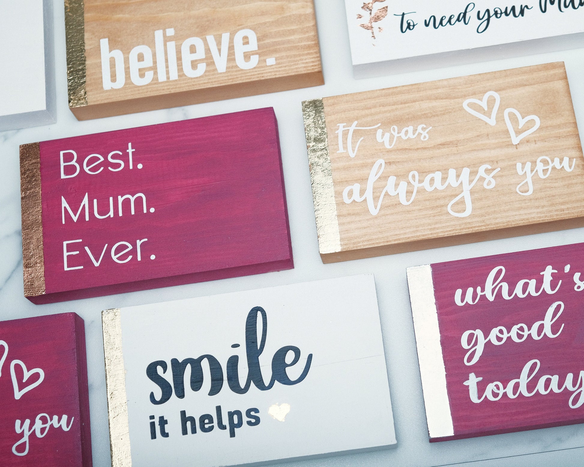 Eight small wooden signs in different colors and with different quotes.  Displayed as tiles on a white desk background, with some signs not fully in the photo. Photo taken from above looking down. Arranged to show other products offered by shop.