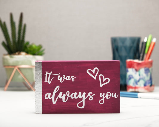 Small freestanding rectangular wooden sign facing straight on. Magenta with silver vertical border on left side. White painted message, It was always you, with two heart outlines top right corner. Plant and pen pot out of focus in background.