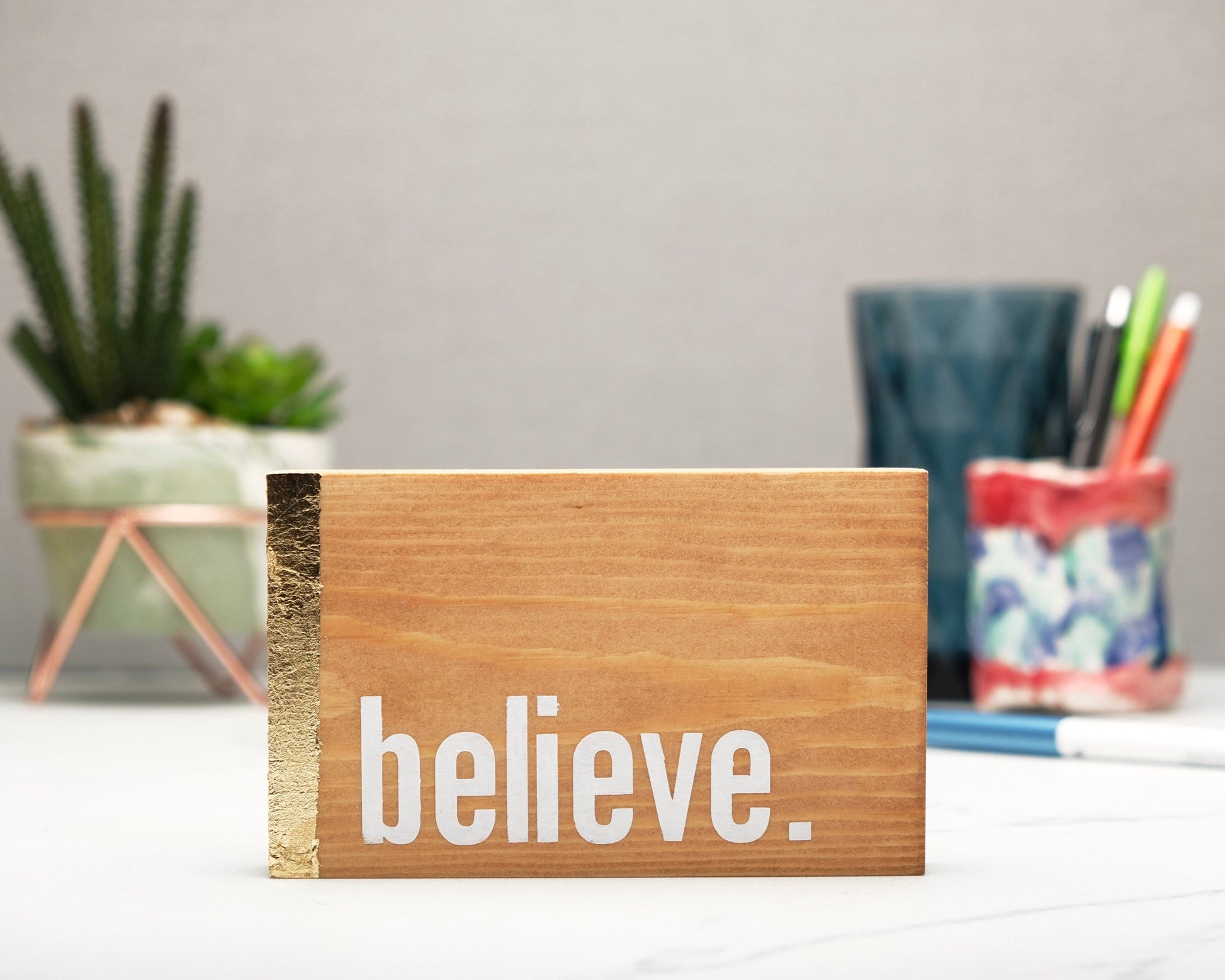 Small freestanding rectangular wooden sign facing straight on. Natural wood color with gold vertical border on left side. White painted message in chunky font with full stop, believe. Plant and pen pot out of focus in background.