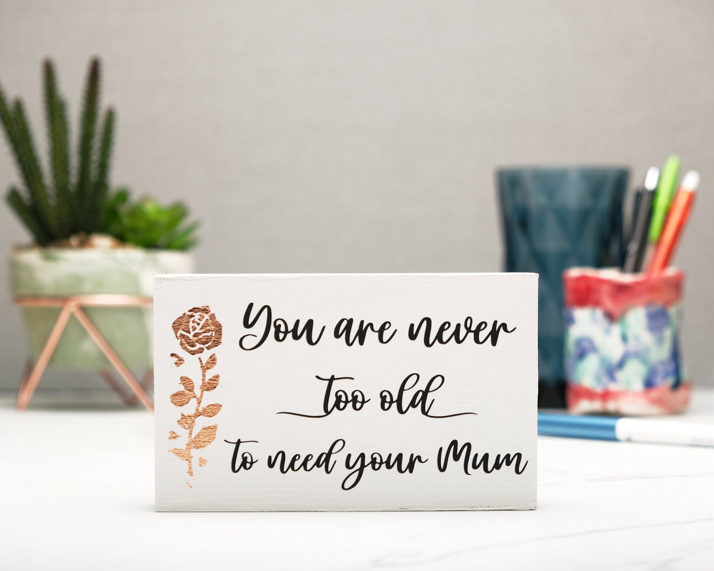 Small freestanding rectangular wooden sign facing straight on. White wood stain with bronze rose gold tall rose flower on left side. Black message in emotive font, You are never too old to need your Mum. Plant and pen pot out of focus in background.