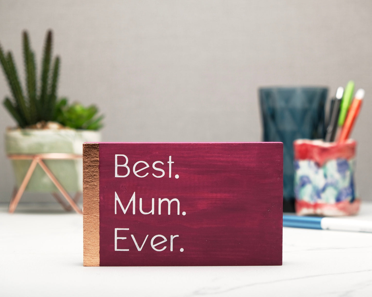 Small freestanding rectangular wooden sign facing straight on. Magenta color, bronze rose gold vertical border on left side. White painted quote in thin font, Best. Mum. Ever. Plant and pen pot out of focus in background.