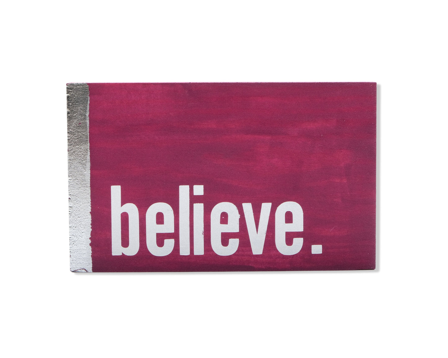Small freestanding rectangular wooden sign facing straight on. Magenta wood stain with silver vertical border on left side. White painted message in chunky font with full stop, believe. Studio style photo with white background.