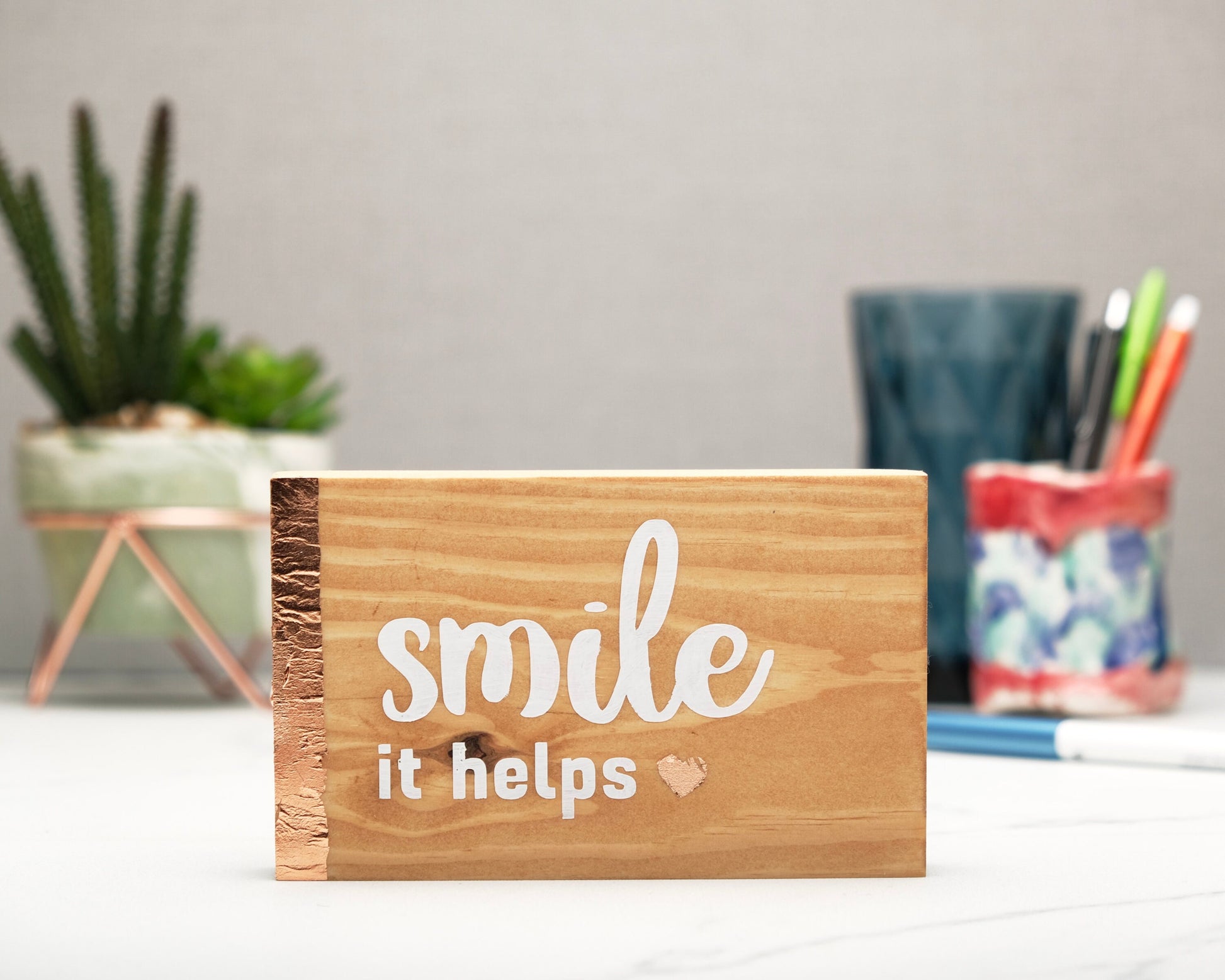 Small freestanding rectangular wooden sign facing straight on. Natural wood color, bronze rose gold vertical border on left side. White painted quote, Smile it helps. Small solid bronze rose gold heart. Plant and pen pot out of focus in background.