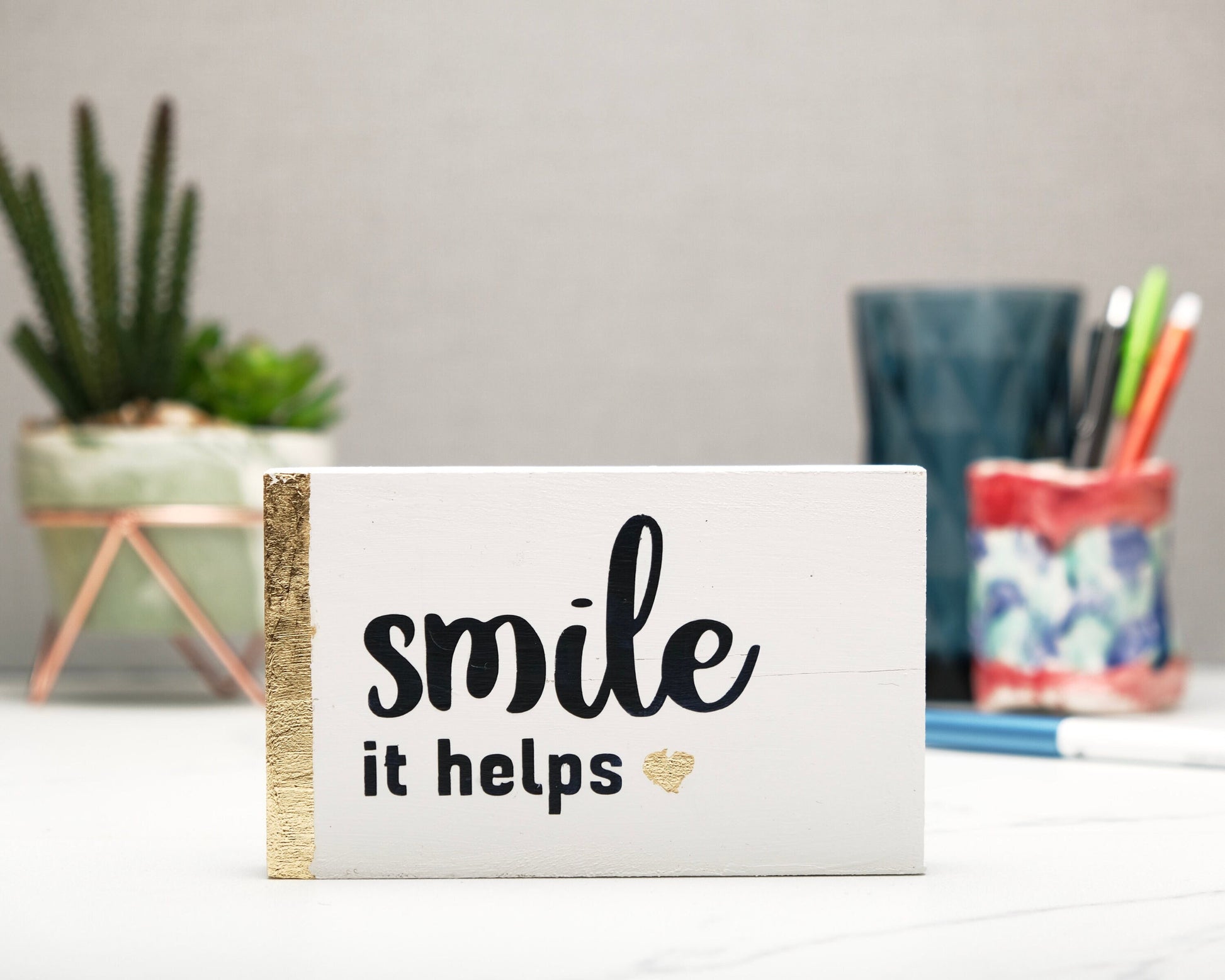 Small freestanding rectangular wooden sign facing straight on. White wood stain, gold vertical border on left side. Black painted quote, Smile it helps. Small solid bronze rose gold heart. Plant and pen pot out of focus in background.