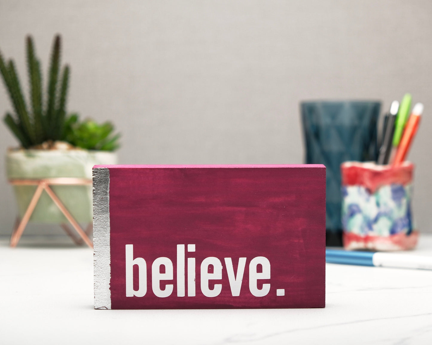 Small freestanding rectangular wooden sign facing straight on. Magenta wood stain with silver vertical border on left side. White painted message in chunky font with full stop, believe. Plant and pen pot out of focus in background.
