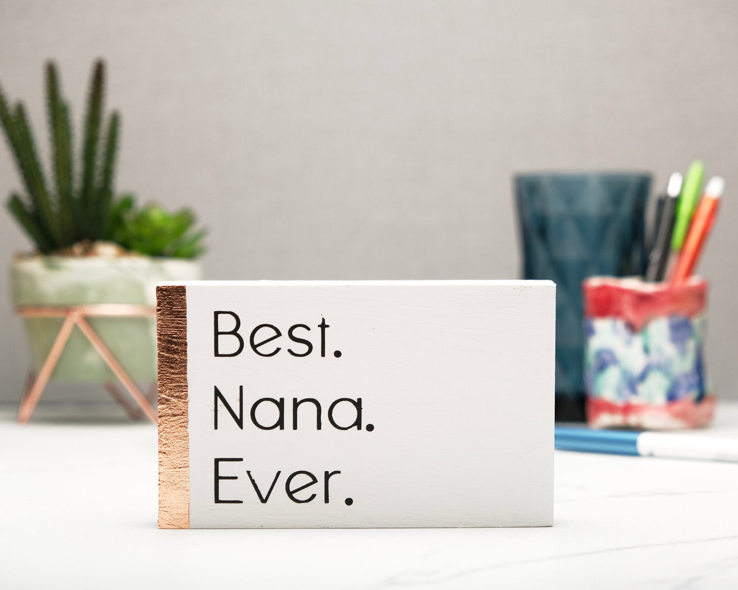 Small freestanding rectangular wooden sign facing straight on. White color with bronze rose gold leaf vertical border on left side. Black painted message in thin font, Best. Nana. Ever. Plant and pen pot out of focus in background.
